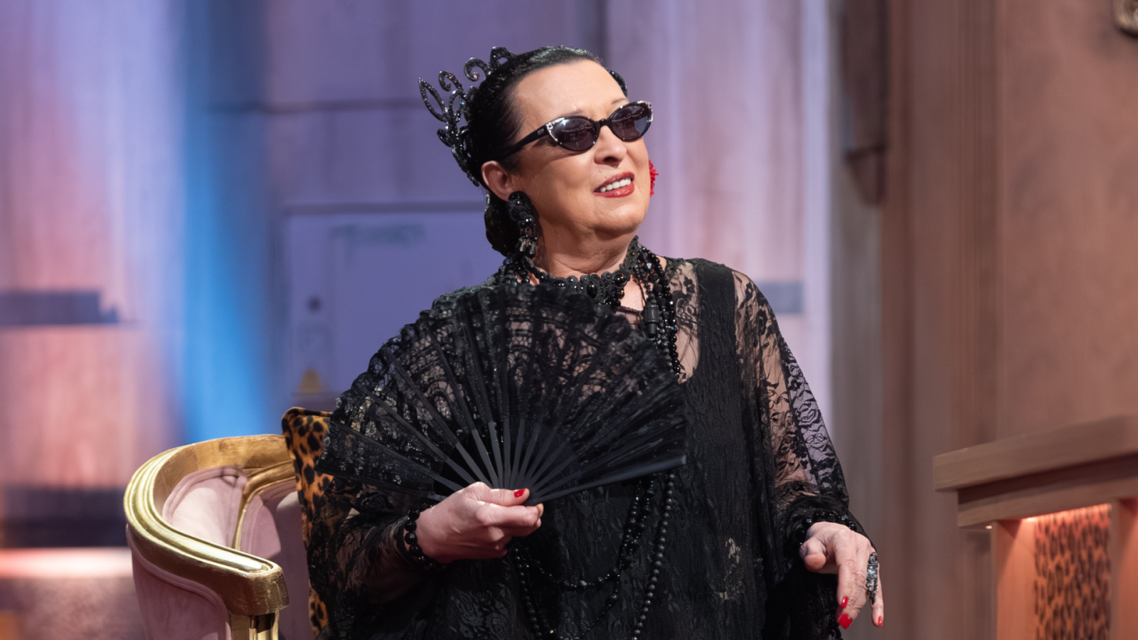 Martirio, artista española, posa con su distintivo estilo: peineta negra, gafas de sol, encaje, abanico y collares. Sentada en un sillón clásico, sonríe con mirada hacia arriba.