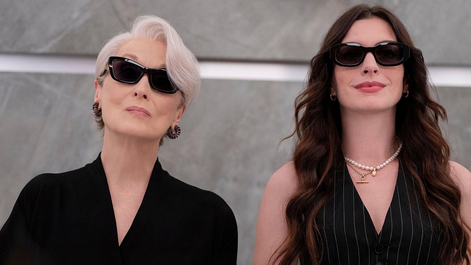 Primer plano de Meryl Streep y Anne Hathaway, ambas con gafas de sol y atuendos oscuros. Streep con cabello corto y canoso, Hathaway con cabello largo y castaño, luciendo collares y pendientes.