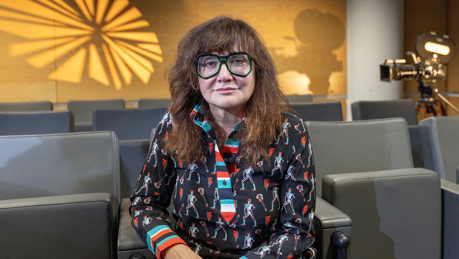 Isabel Coixet, directora de cine, sentada en un auditorio con gafas y camisa estampada. Al fondo, butacas y una cámara de cine antigua.