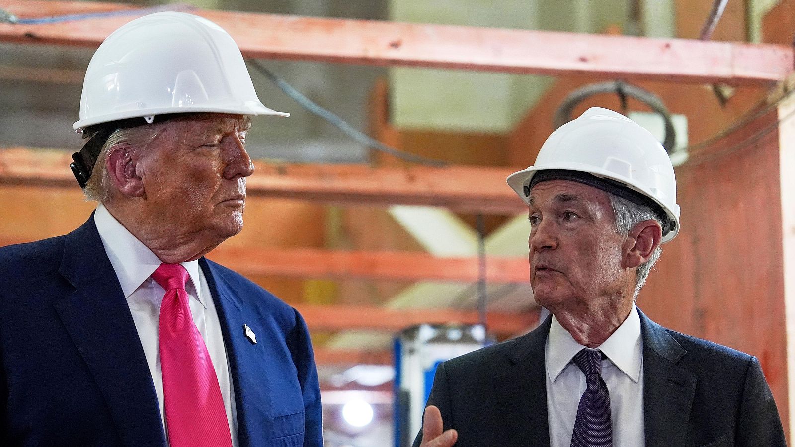 Donald Trump y Jerome Powell, con cascos y trajes, conversan en un interior industrial. Powell gesticula mientras Trump escucha atentamente.
