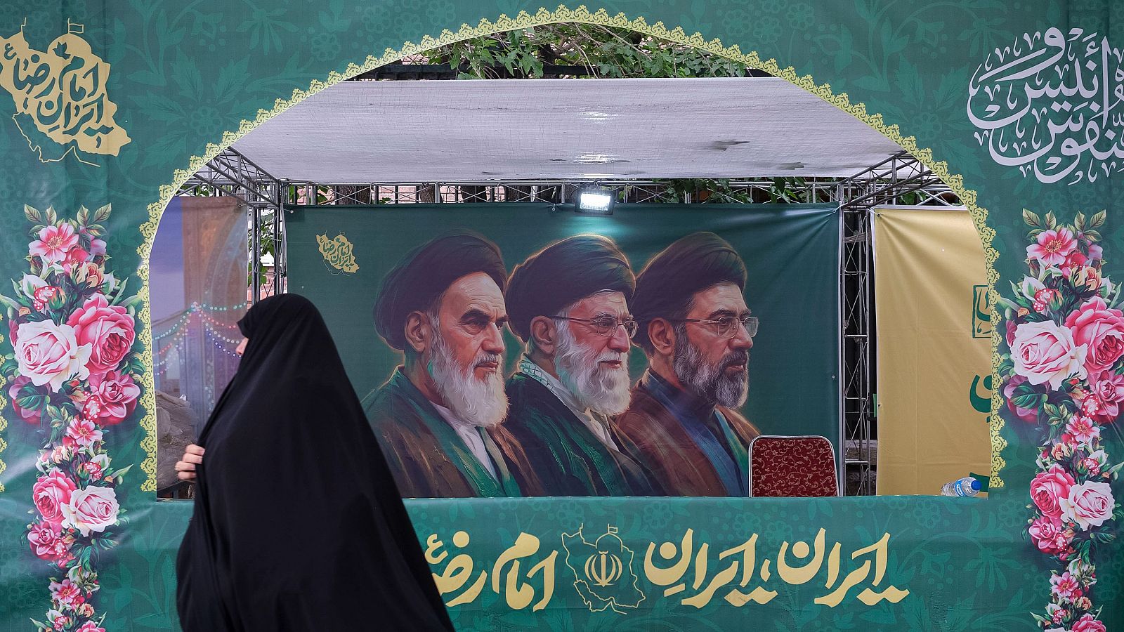 Una mujer vestida con el tradicional chador pasa delante de un cartel en Teherán que muestra a los tres líderes supremos de la República Islámica, incluido el actual Mojtaba Jameneí. Miles