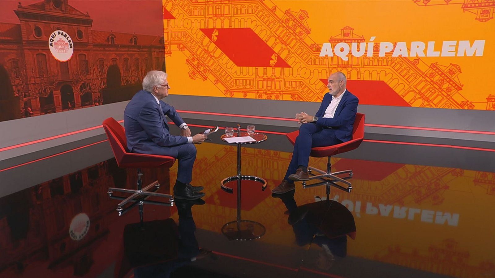 Lluís Falgàs entrevista a Joan Ridao en el programa "AQUÍ PARLEM", con paneles digitales de fondo y un suelo reflectante.