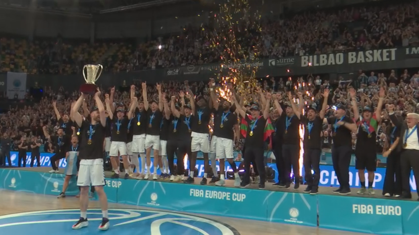 Jugadores del Surne Bilbao Basket celebran la victoria de la FIBA Europe Cup en un podio, alzando el trofeo dorado. Confeti y chispas doradas caen sobre ellos, mientras el público anima desde las gradas.