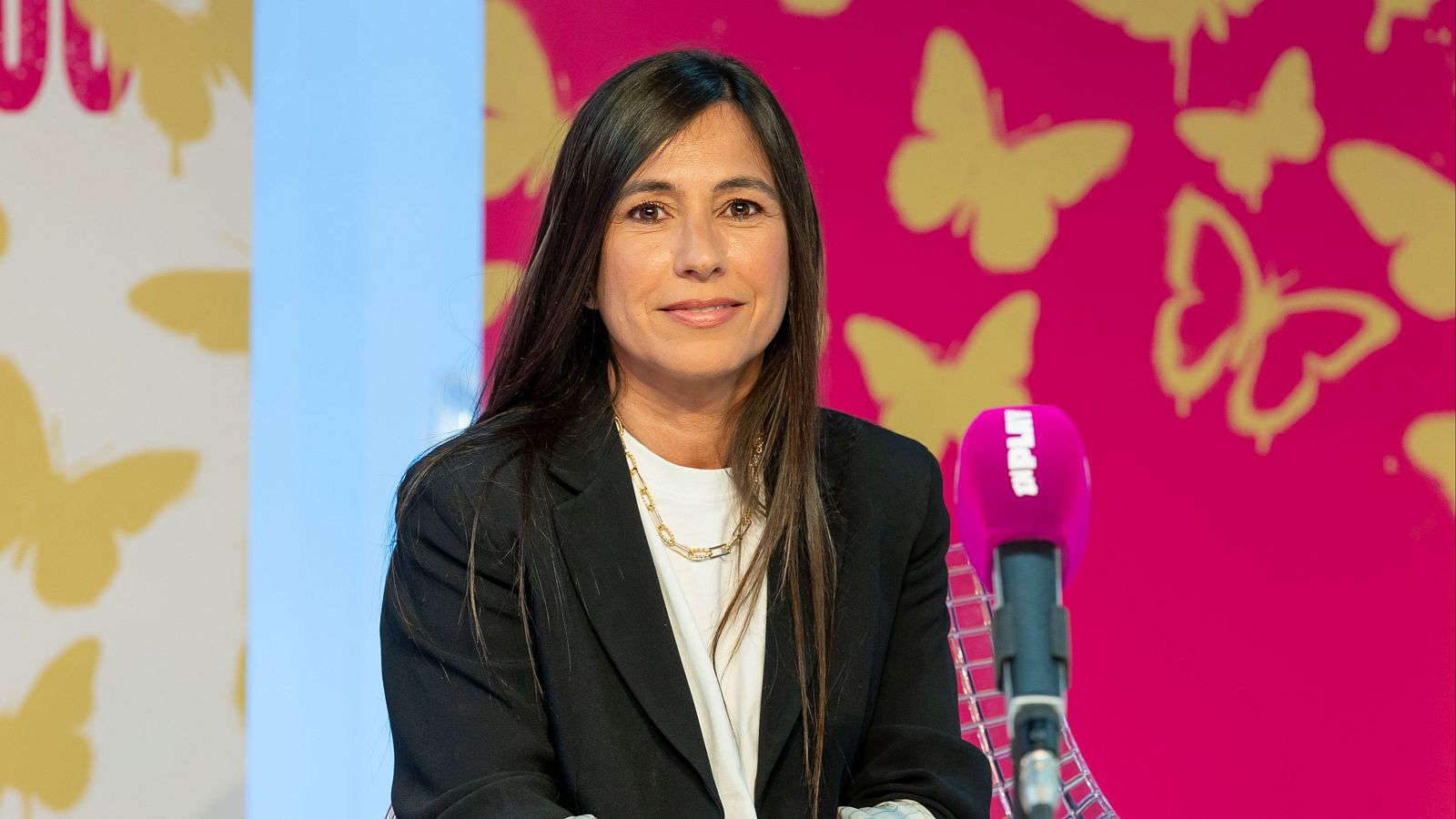 Ana Gómez, ex directiva de plataformas digitales, sonríe a cámara con un collar dorado y blazer negro. Un micrófono fucsia con "AMPLIAR" se ve a su lado. Fondo blanco y fucsia con mariposas.