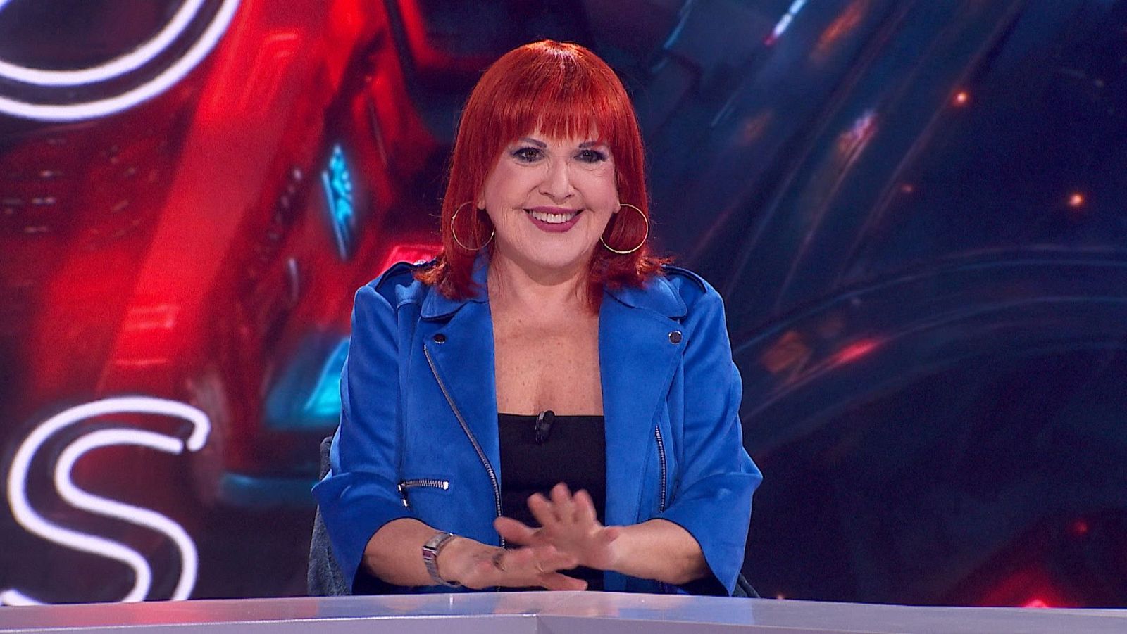 Loles León, actriz española, posa en un estudio de televisión con chaqueta azul eléctrico y cabello rojo anaranjado. Fondo con luces rojas y azules y una gran letra 'S'.