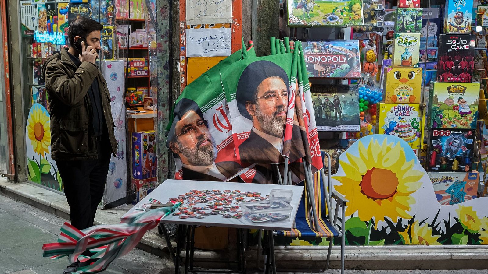 Calle nocturna en Teherán con un hombre hablando por teléfono junto a un puesto de pines y banderas de Ebrahim Raisi. Escaparate de tienda con juegos de mesa al fondo.
