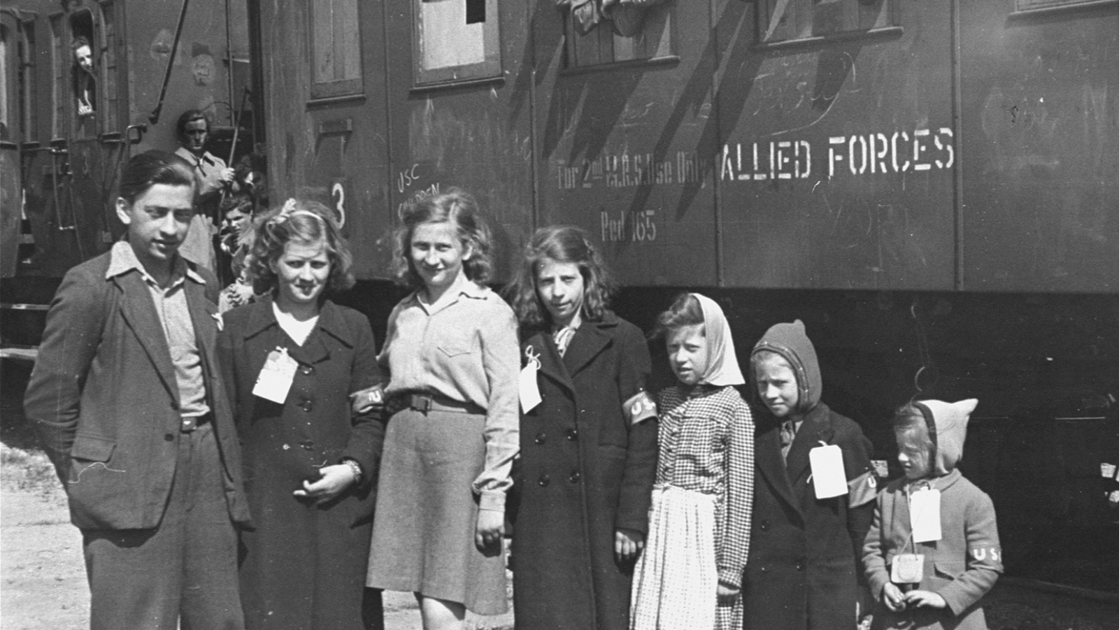 Grupo de siete personas, un joven y seis niños, con etiquetas y brazaletes "USC", frente a un vagón de tren "ALLIED FORCES" en Bremerhaven, Alemania, el 11 de mayo de 1946.