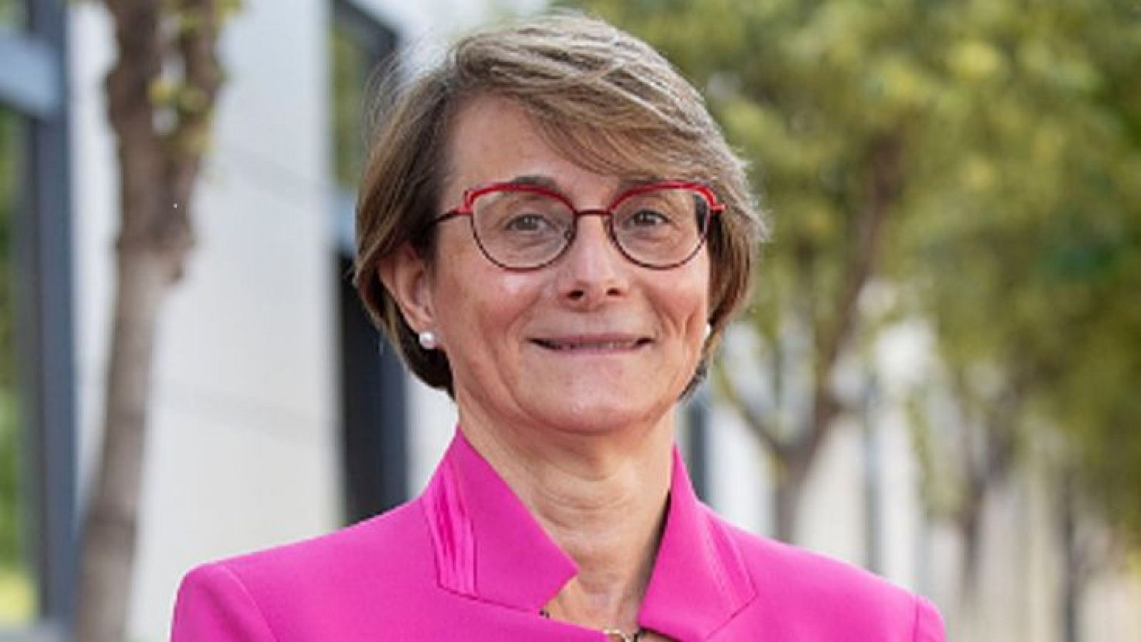 Retrato de Eva Alcón, rectora de la UJI, con gafas rojas y chaqueta fucsia, sonriendo a la cámara.