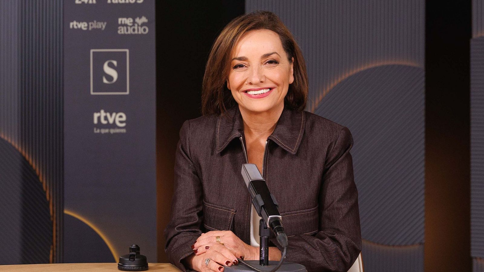 Pepa Bueno, periodista, sonríe en un estudio de grabación con micrófono y logos de RTVE al fondo.