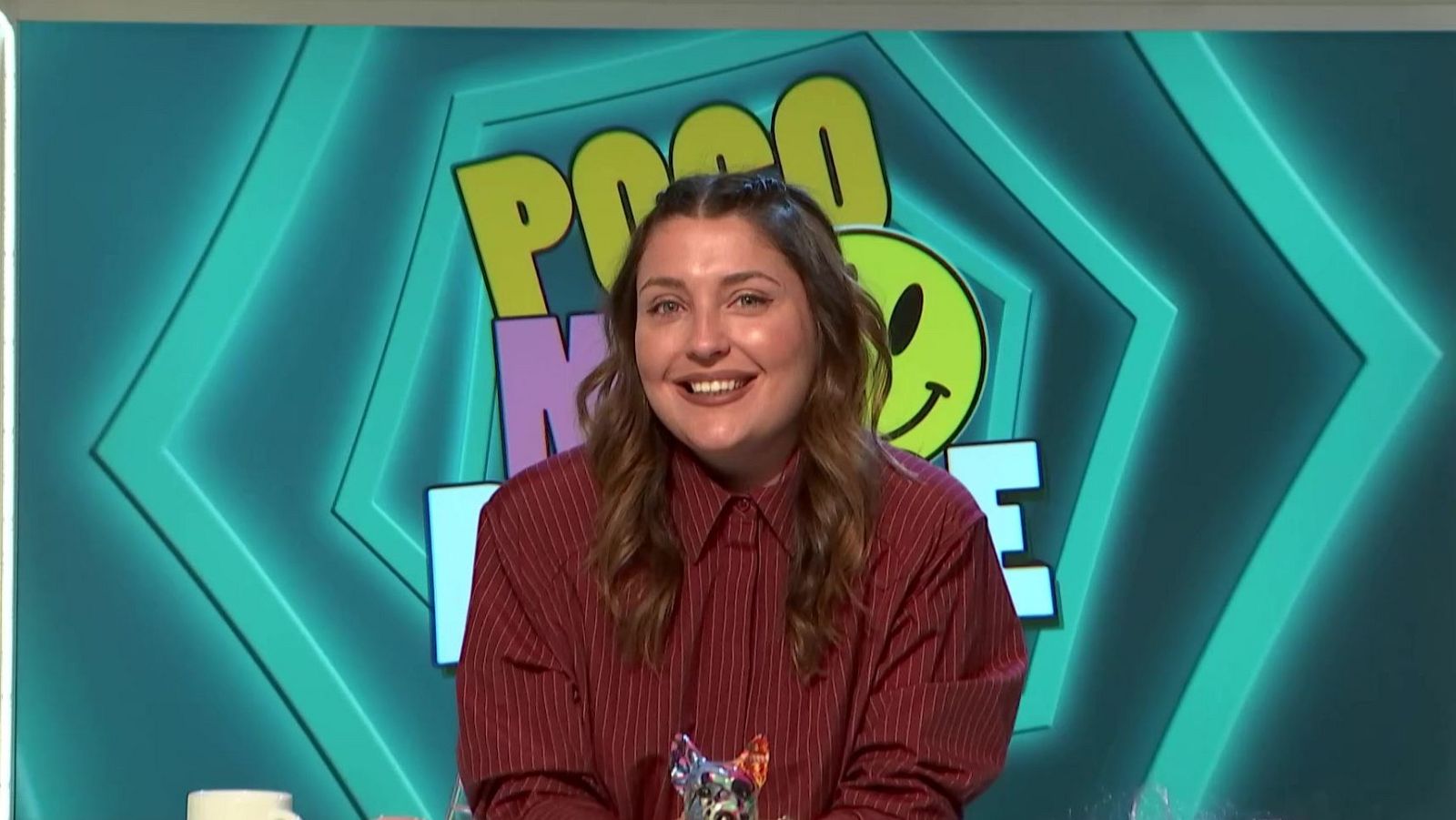 Marina Lobo sonríe en un plató con el logo "POCO ME" y un emoticono verde. Viste camisa roja y su pelo es castaño y liso.