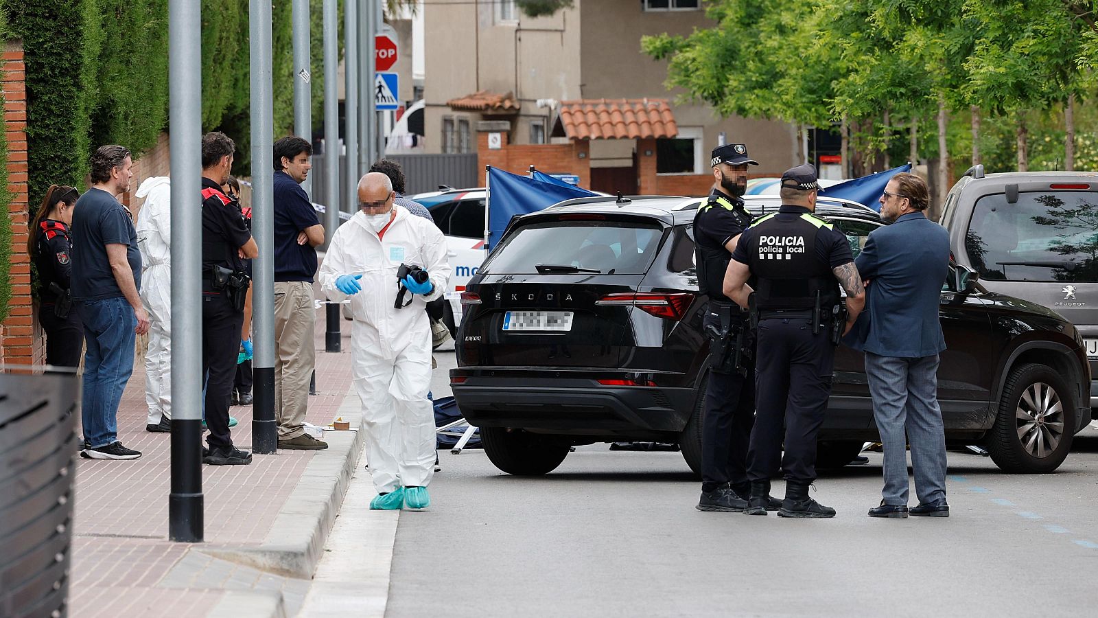 Investigador forense con traje blanco y guantes azules examina pruebas en acera. Policías y hombre con traje observan escena junto a vehículos. Área acordonada con lonas azules.