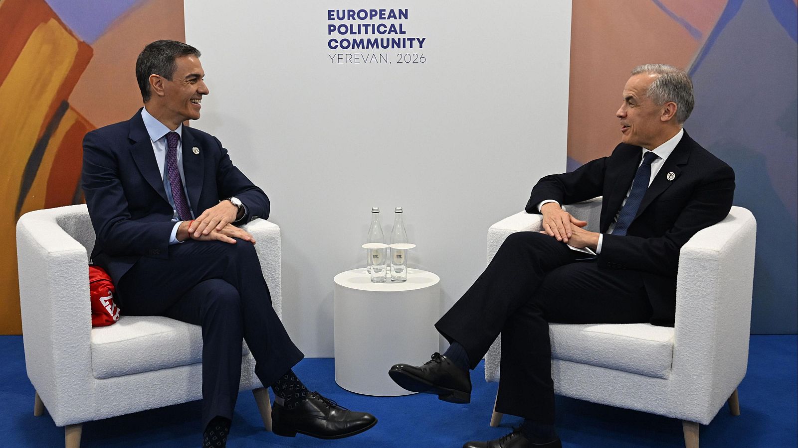 Macron y Pashinyan en la Cumbre de la Comunidad Política Europea en Ereván 2023. Pashinyan señala mientras habla. Fondo blanco con logo de la Comunidad Política Europea.