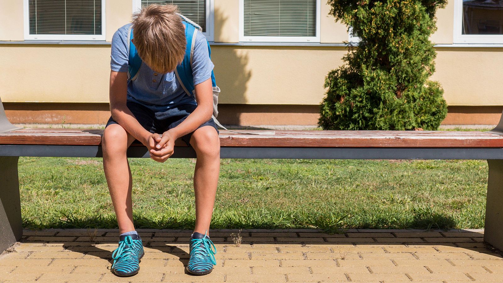 Fotografía de un niño Infeliz niño triste sentado en posición cerrada con la cabeza baja sentado en un banco del patio de un colegio