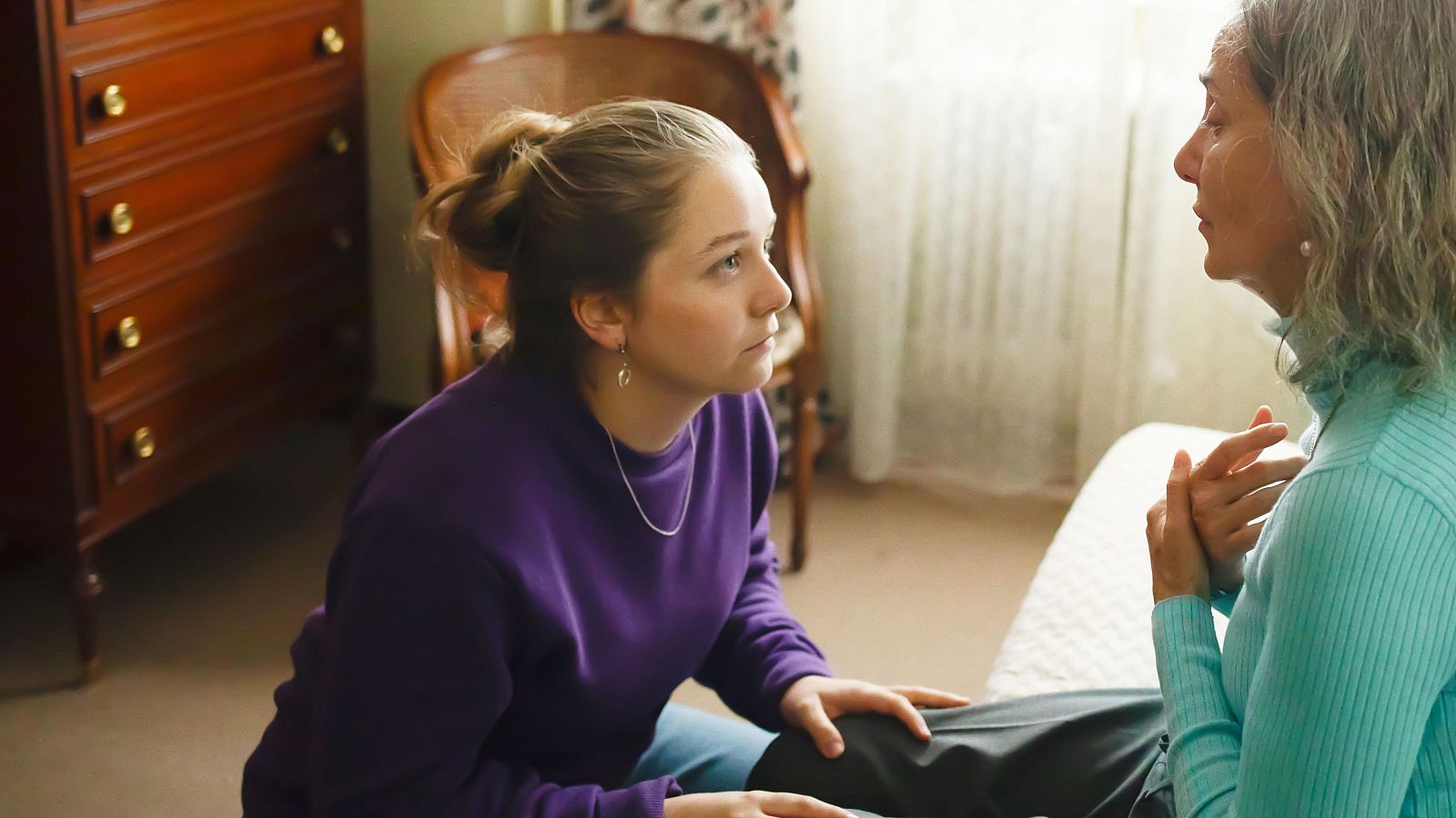 Olivia Delcán y Emma Suárez comparten un momento íntimo en una habitación luminosa. Olivia, de rubio y sudadera morada, se arrodilla ante Emma, de pelo canoso y jersey azul turquesa.