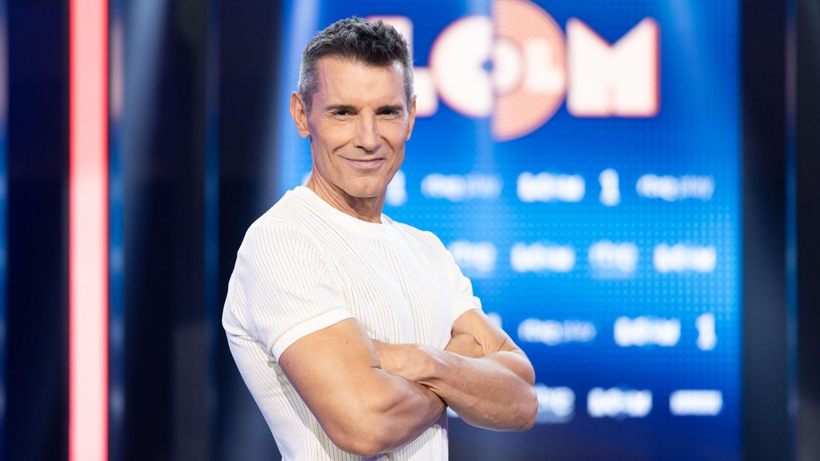 Jesús Vázquez sonríe a cámara con camiseta blanca de rayas, sobre un fondo azul con "COL", "RTVE" y "La 1".