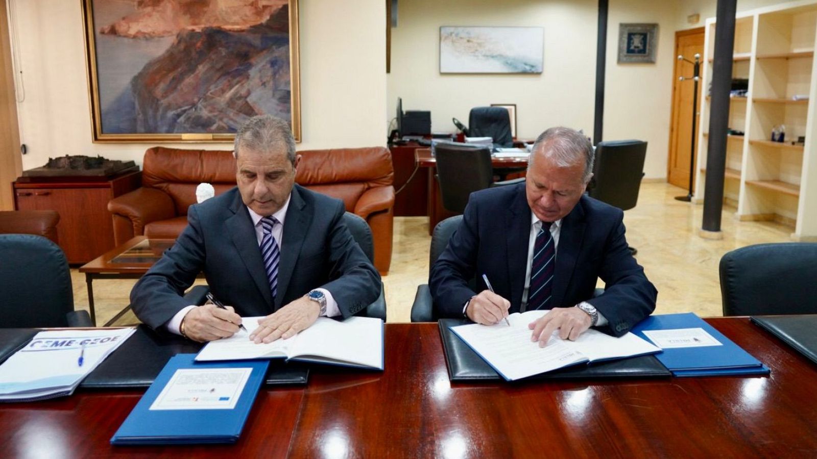 Enrique Alcoba y Miguel Marín firman convenios de colaboración en una oficina. Ambos visten trajes oscuros y corbatas, concentrados en la firma de documentos sobre una mesa de madera.