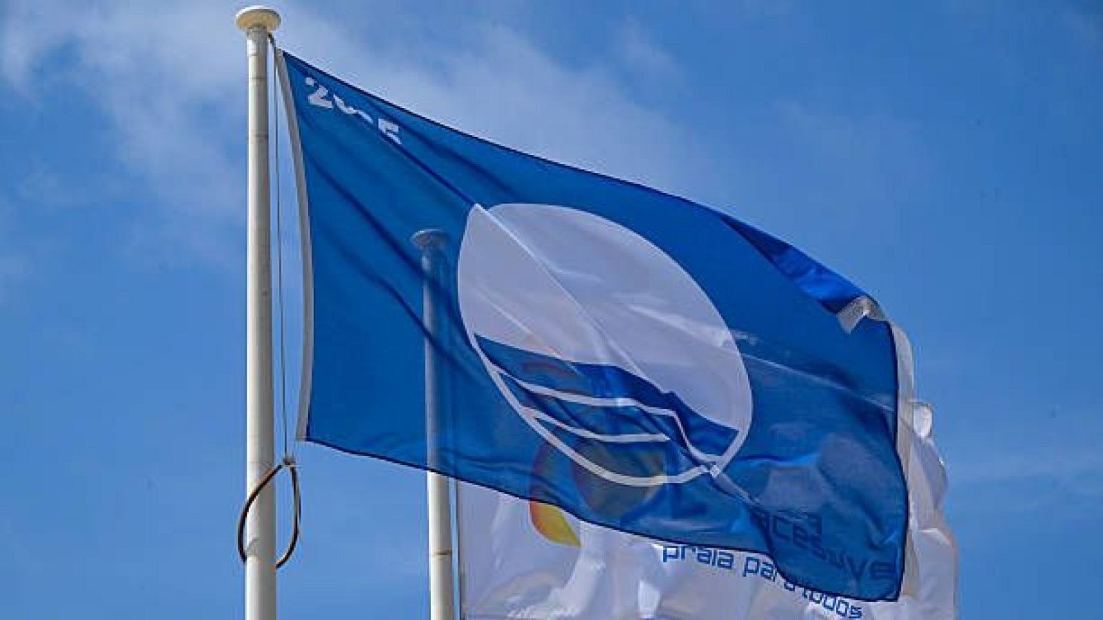 Bandera Azul con "2023" y otra bandera con "praia para todos" y "acessível" ondean en un mástil blanco bajo cielo azul.