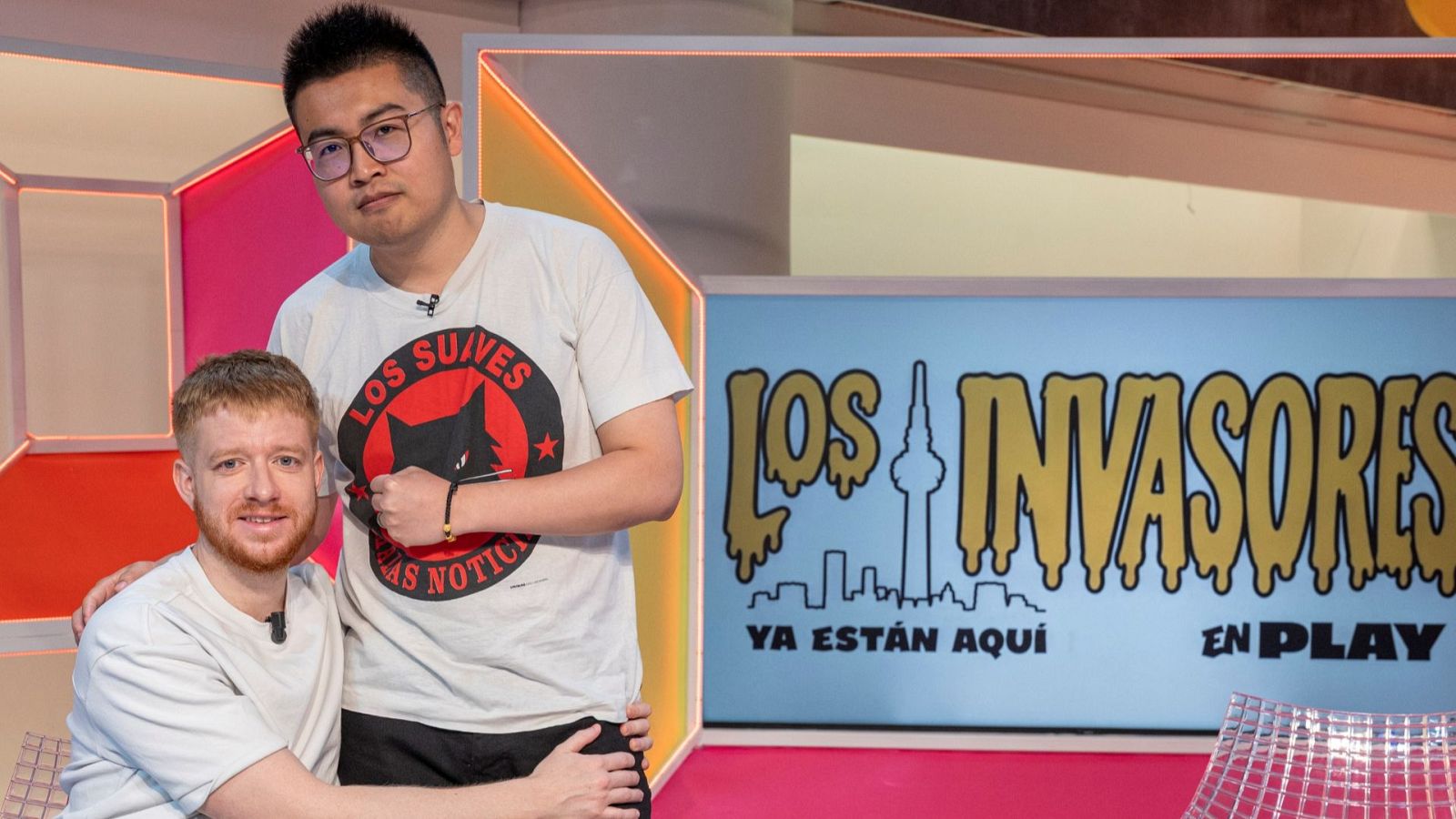 Alex Ayres y Jiajun Yin, presentadores de "Los invasores", posan en un estudio de televisión. Jiajun lleva una camiseta con el logo "LOS SUIVES LAS NOTICIAS" y un gato negro.