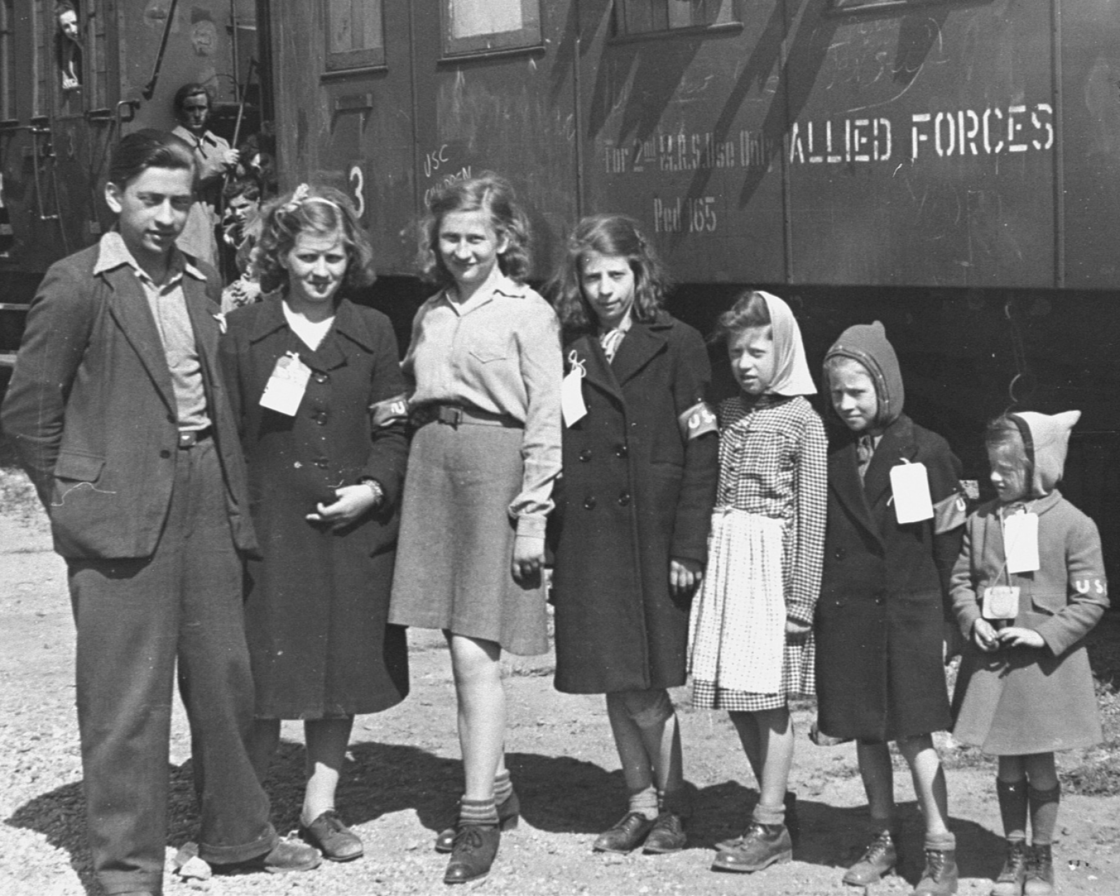 Grupo de siete personas, un joven y seis niños, posan frente a un vagón de tren con inscripciones como "ALLIED FORCES". Los niños portan distintivos y bandas, indicando un posible contexto de posguerra.