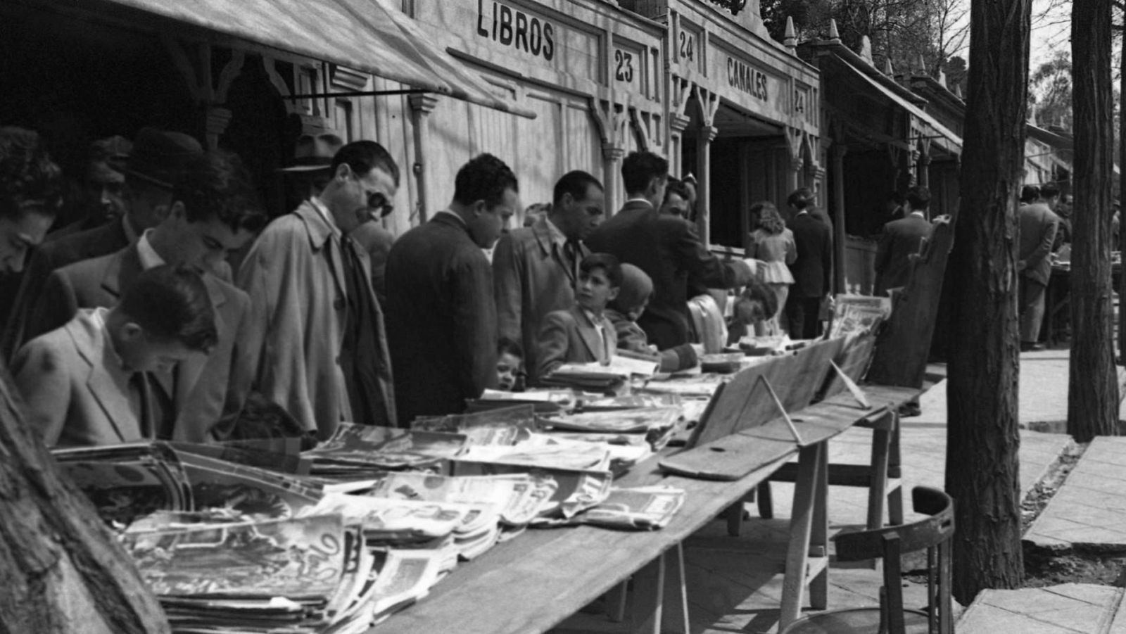 Mercado de libros en la Cuesta de Moyano, Madrid. Puestos de madera con "LIBROS" y números "23", "24", "CANALES". Personas de época examinando libros en mesas.