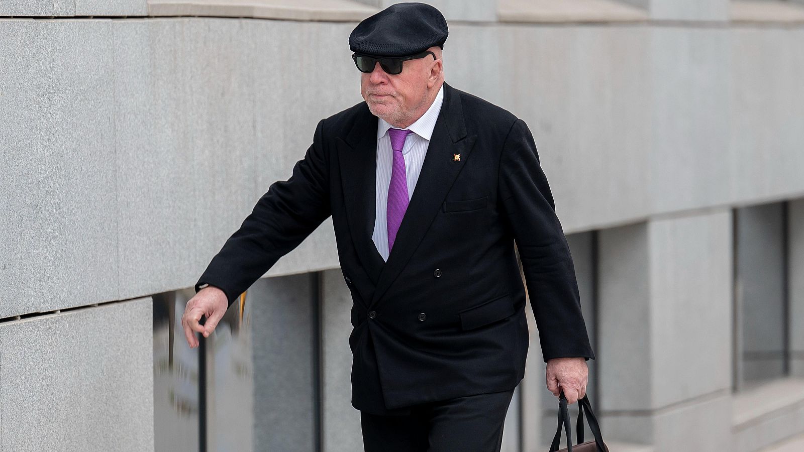 Un hombre con traje negro, camisa blanca y corbata morada, identificado como José Manuel Villarejo, camina llevando un maletín. Lleva gafas de sol y una gorra oscura.