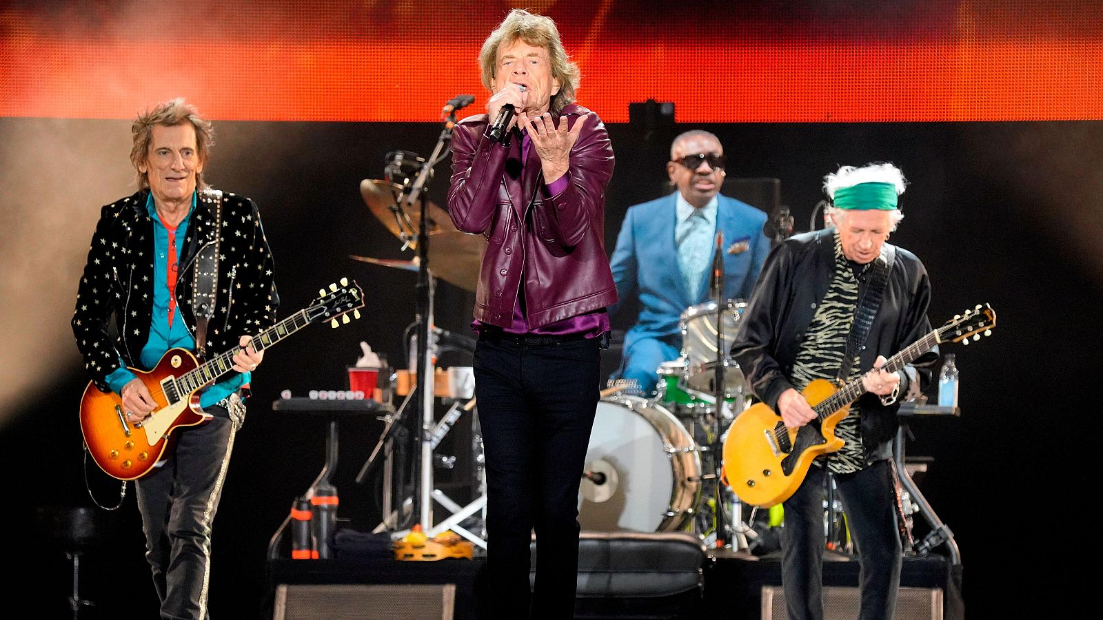 Cuatro miembros de The Rolling Stones en concierto: Ronnie Wood y Keith Richards con guitarras, Mick Jagger cantando y Steve Jordan en la batería. Escenario iluminado con luces rojas.