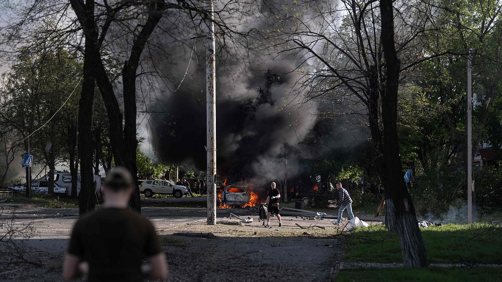 Un coche arde en una calle arbolada, rodeado de escombros y observadores. El humo negro se eleva, creando una atmósfera de destrucción.