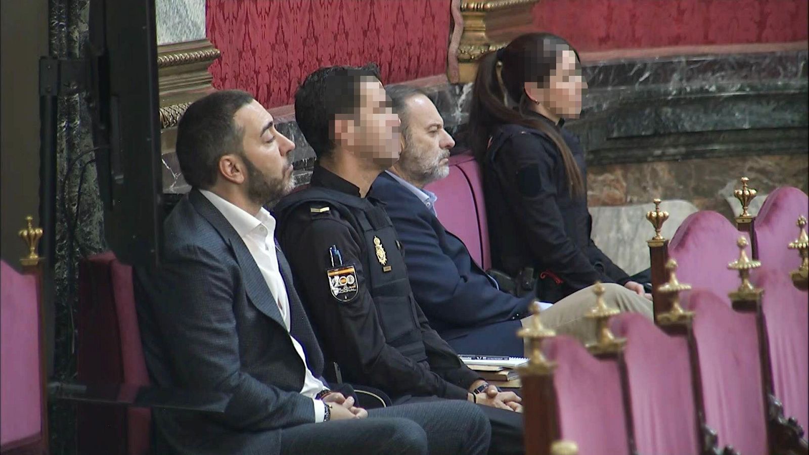 Alberto Luceño en sala de audiencias, flanqueado por Policía Nacional y otras personas. Fondo formal con tela roja y detalles dorados.