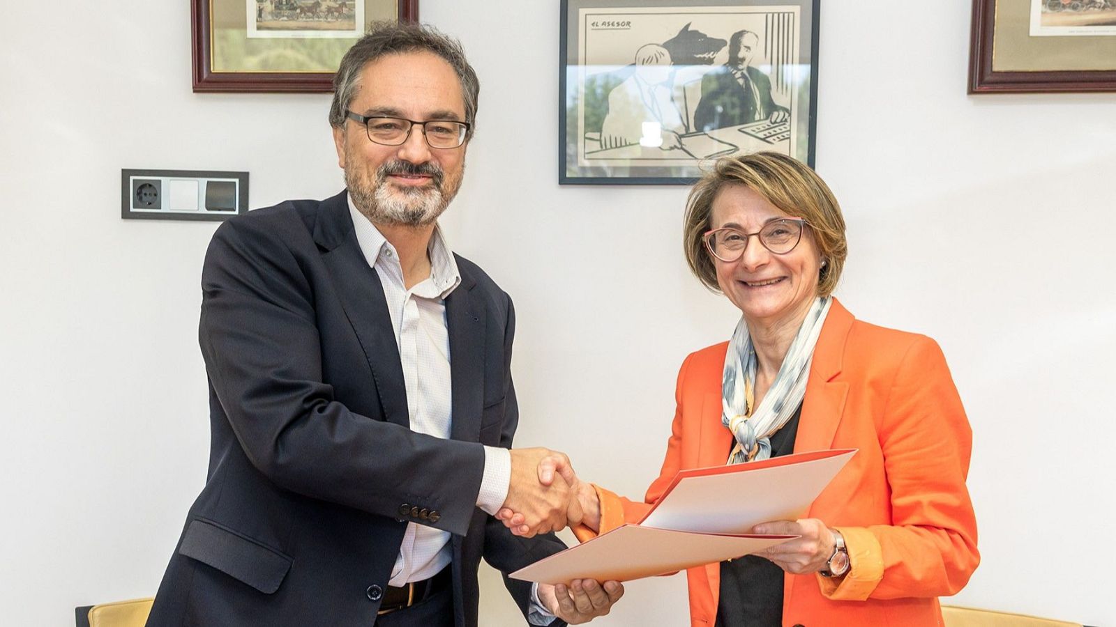 Alfonso Morales y Eva Alcón, sonriendo, se dan la mano en una oficina. Él viste traje oscuro y ella chaqueta naranja. Al fondo, una pared blanca con cuadros y un enchufe.