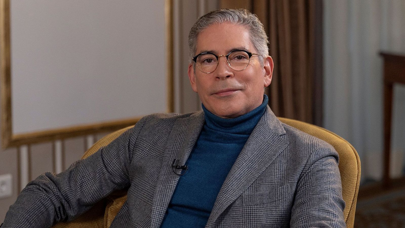 Retrato de Boris Izaguirre, escritor y presentador, con cabello canoso, gafas y jersey azul, sentado en sillón mostaza. Fondo interior desenfocado.
