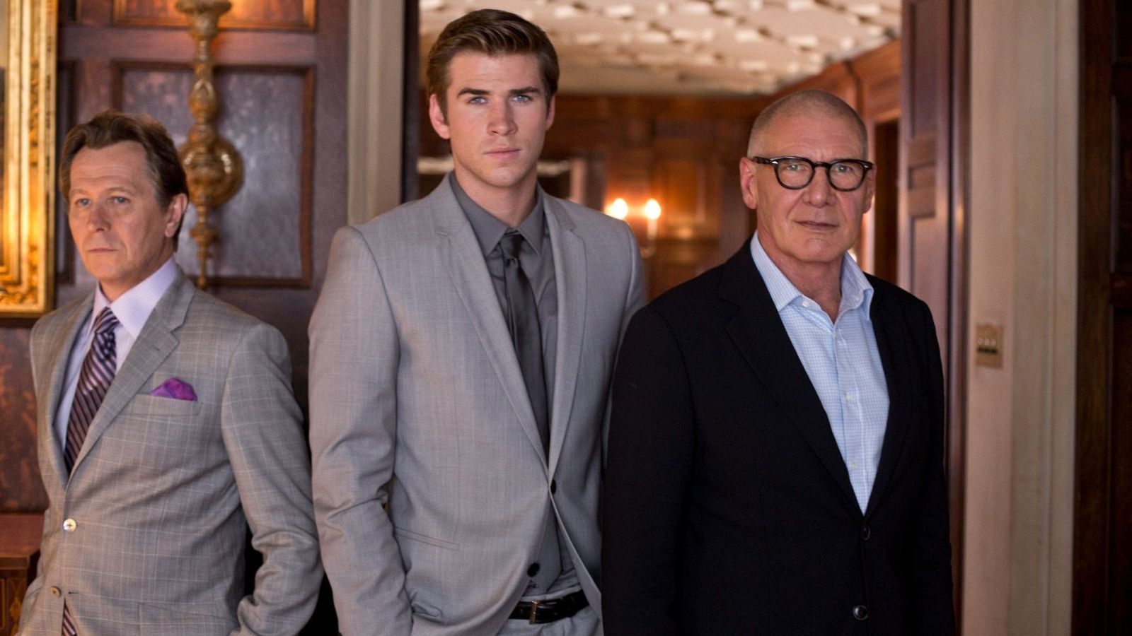 Gary Oldman, Liam Hemsworth y Harrison Ford posan juntos en un interior elegante, vestidos con trajes formales. Cada uno muestra un estilo distinto en sus atuendos y expresiones.