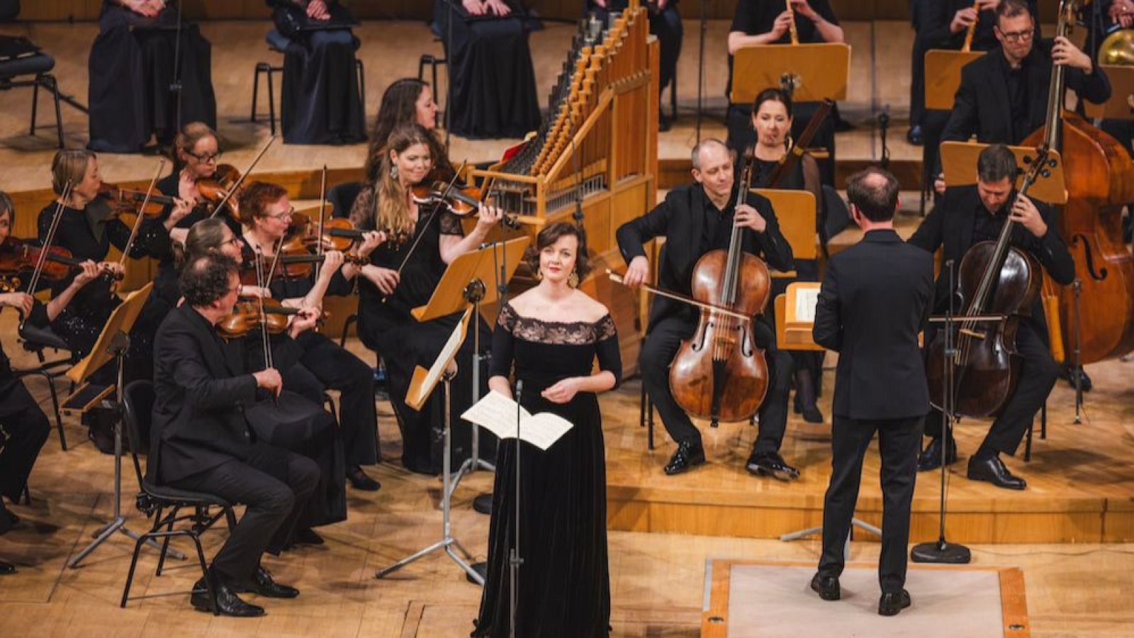 Soprano Maria Ladurner y el director dirigen al conjunto instrumental Barucco en un concierto. Se observan músicos de cuerda, un instrumento de teclado y atriles en un escenario de madera.