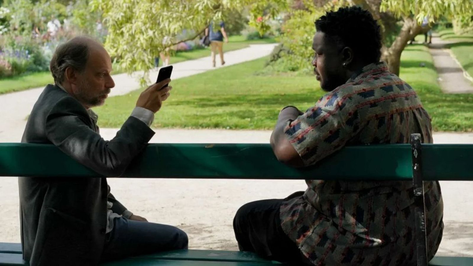 Pierre, absorto en su smartphone, y Baptiste, observándolo, comparten un banco en un parque soleado. Detalle de sus vestimentas y apariencias.