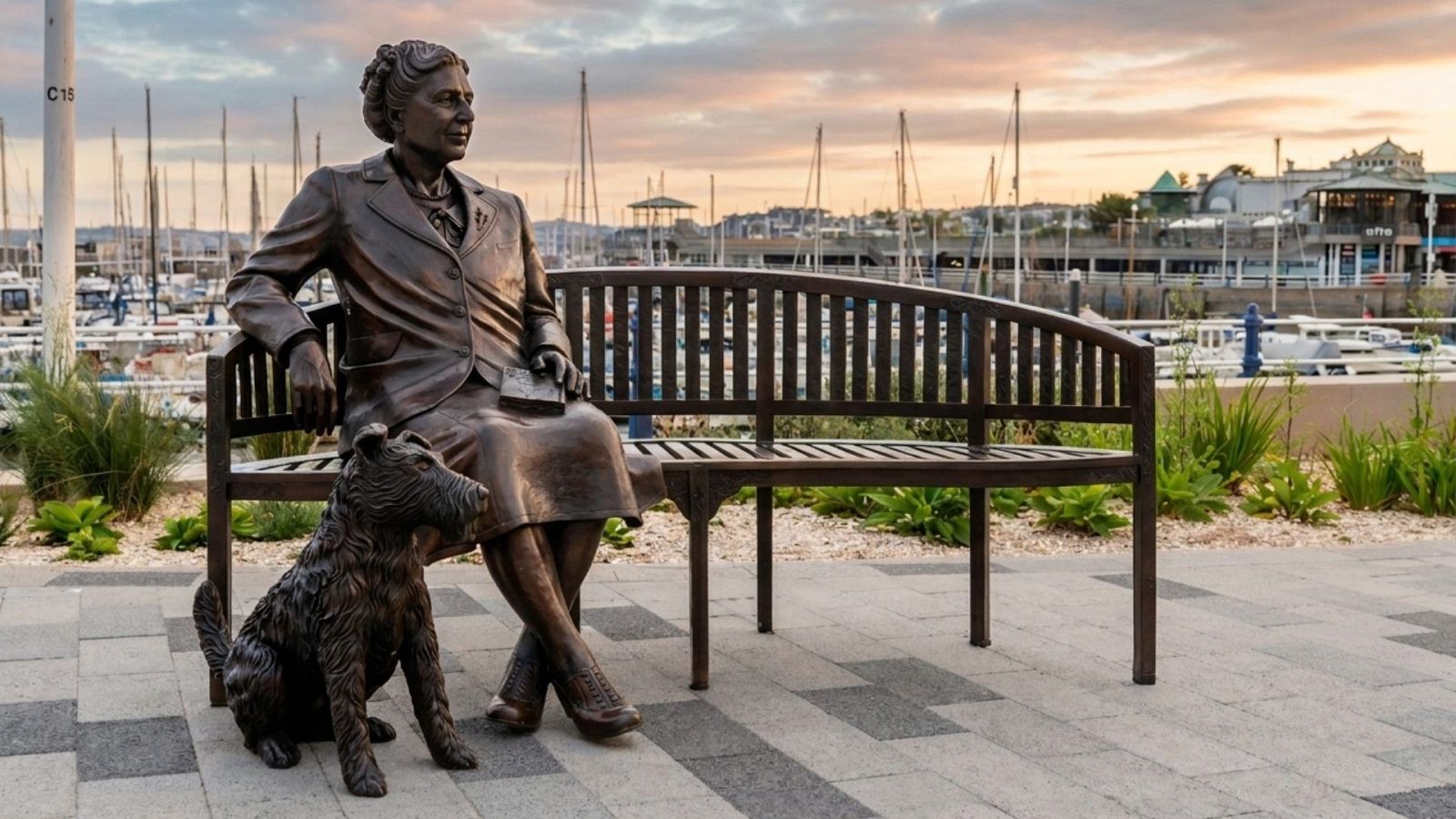 Estatua de Agatha Christie y su perro Peter en un puerto deportivo al atardecer. El cielo muestra tonos naranjas y rosas.