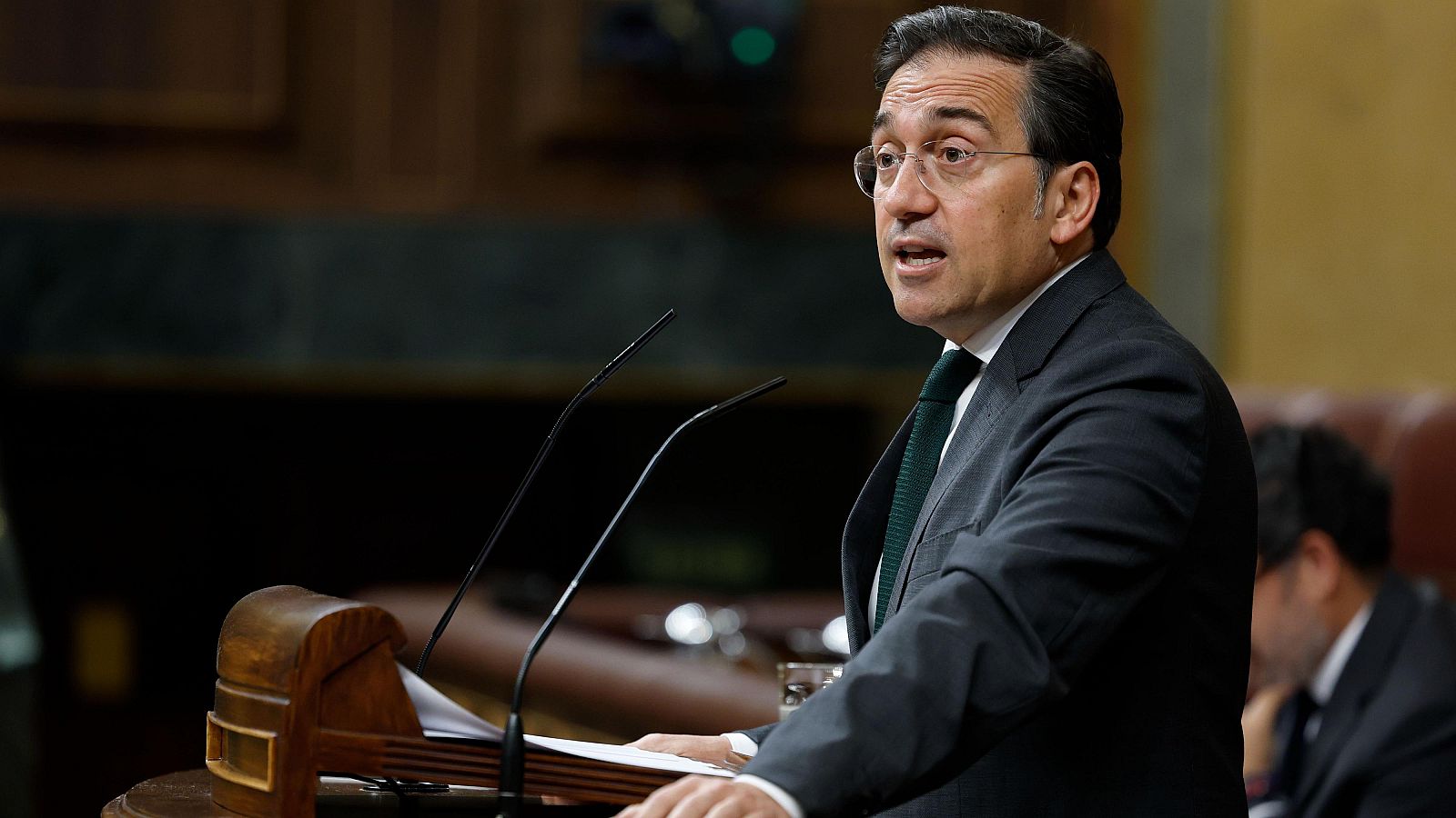 Primer plano de José Manuel Albares, ministro de Asuntos Exteriores, hablando en el Congreso de los Diputados. Lleva gafas, traje oscuro y corbata verde. Sobre el atril, micrófonos y papeles.