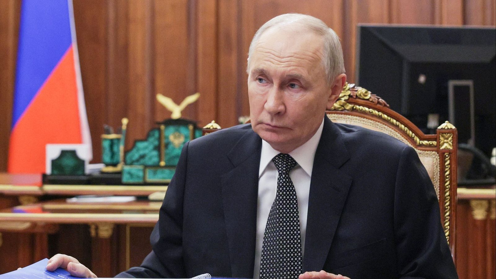 Vladímir Putin, presidente ruso, con traje oscuro y expresión seria, sentado en un entorno formal con la bandera de Rusia y un águila bicéfala dorada al fondo.