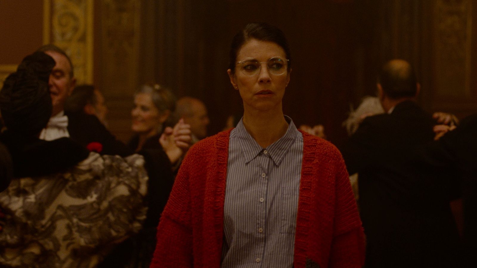 Malena Alterio, con gafas y cárdigan rojo, mira fijamente al frente en un primer plano. Detrás, un salón elegante con gente socializando y bailando.