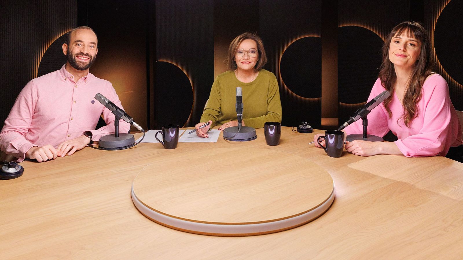 Antón Gómez-Escolar, Pepa Bueno y Paloma Torrecillas posan sonrientes en un estudio, con micrófonos y tazas, ante un fondo de círculos dorados.