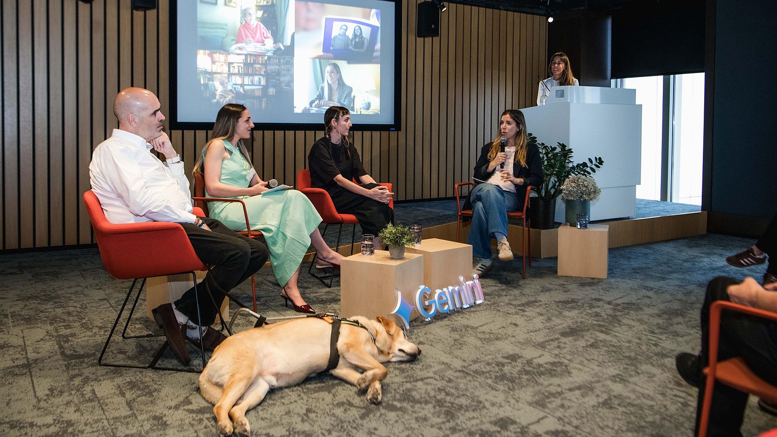Cuatro personas sentadas comparten escenario. En primer término un perro está tumbado. Se pueden leer la palabra Gemini, la IA de Google. En la pared hay una imagen proyectada donde se ve a algunos de los protagonistas que están en el escenario