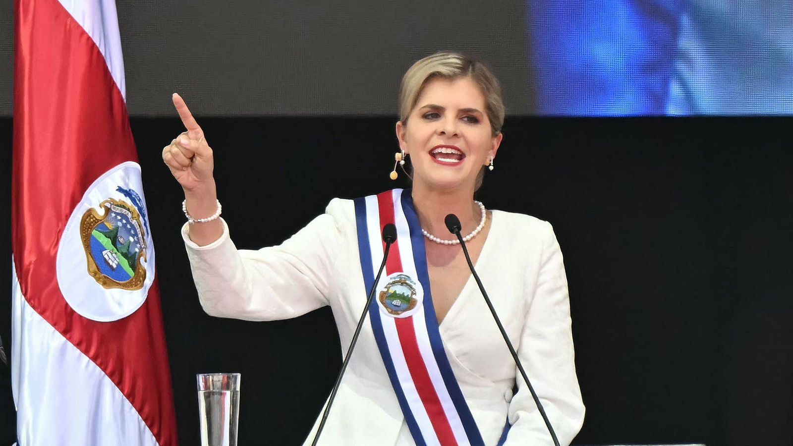 Laura Fernández, presidenta de Costa Rica, en su investidura, habla con la boca abierta y el dedo índice levantado, portando la banda presidencial sobre una blusa blanca y joyas de perlas.