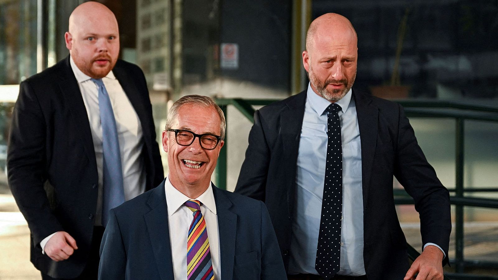 Nigel Farage, líder de Reform UK, sonríe ampliamente con traje azul y corbata colorida, flanqueado por dos hombres serios con traje y barba. Fondo arquitectónico.