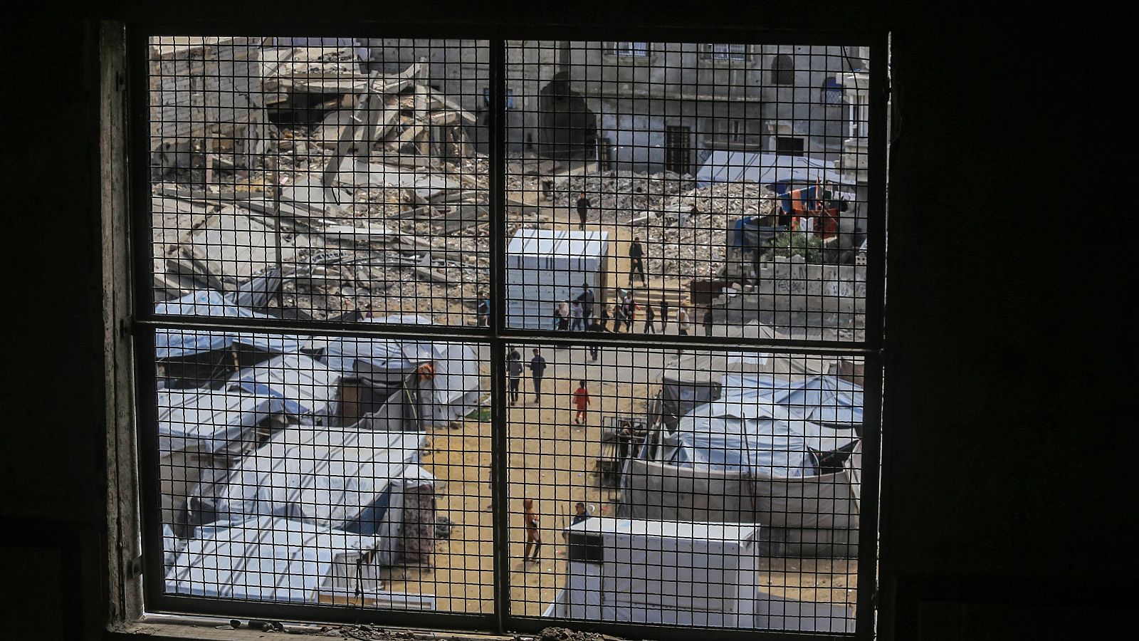 Vista de un campamento de tiendas en Khan Younis, 5 de mayo de 2026, a través de una ventana con reja, mostrando edificios destruidos y personas.