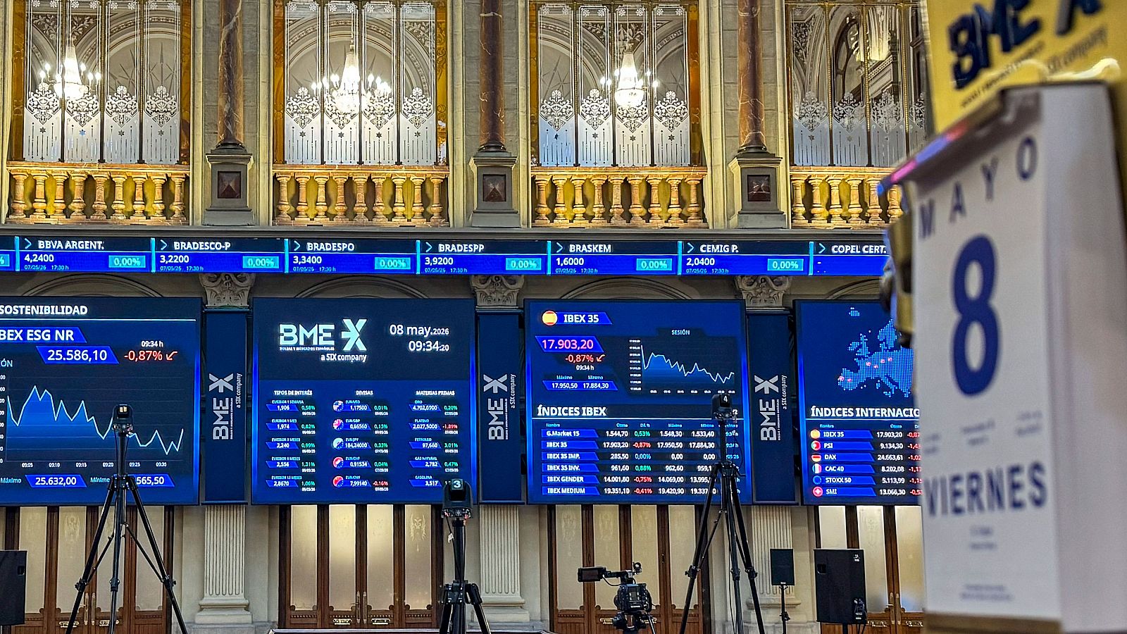Interior de la Bolsa de Madrid (BME X) el 8 de mayo de 2020, con pantallas mostrando cotizaciones a la baja del IBEX 35 y otros índices, y un calendario físico.