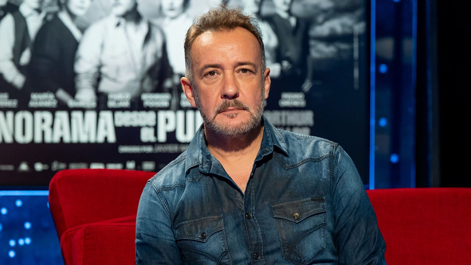 Primer plano de José Luis García-Pérez en sofá rojo, camisa vaquera, barba. Fondo oscuro con pantalla B/N y texto: "NORAMA", "DESDE EL PU".
