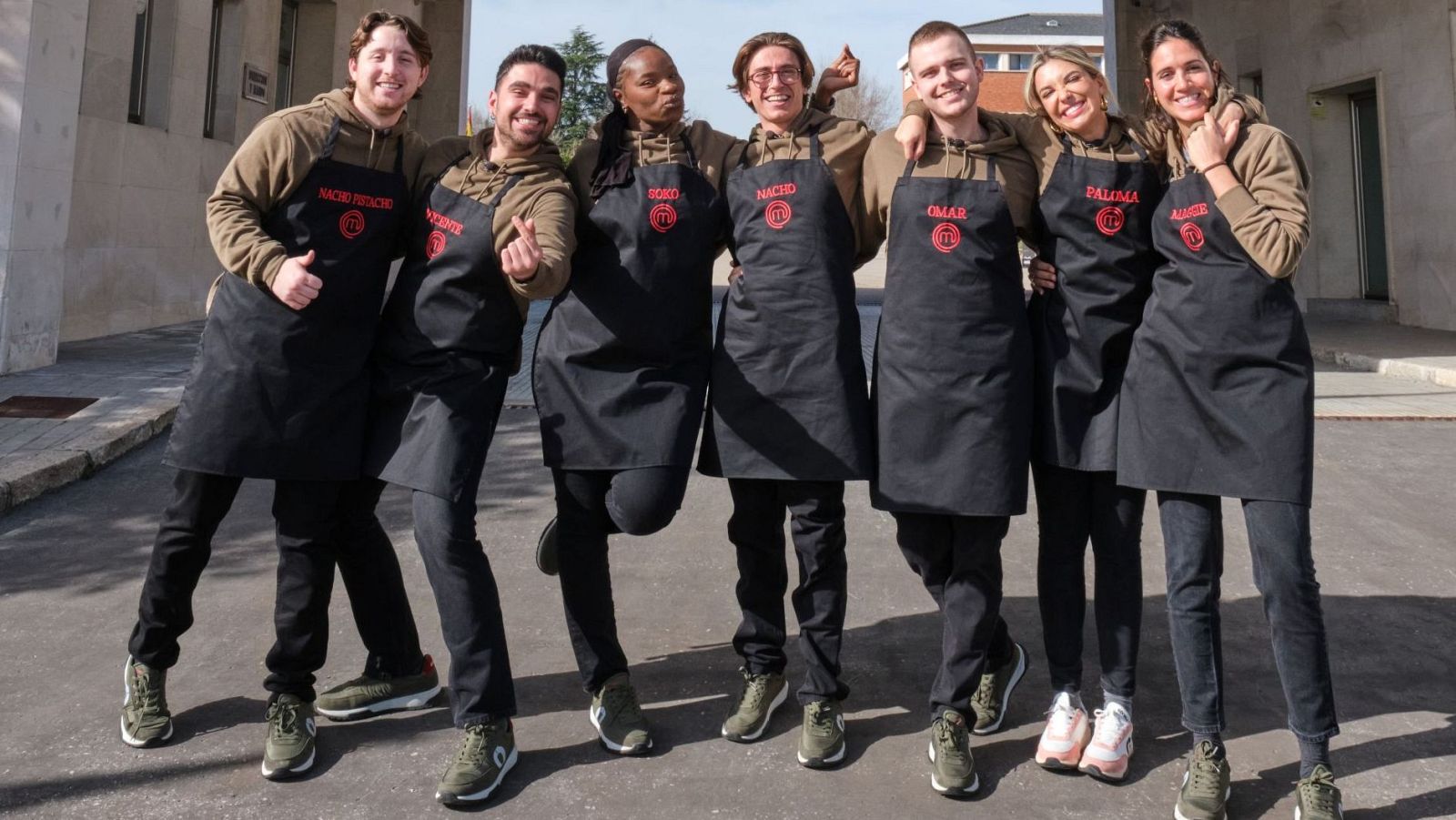 Siete aspirantes de MasterChef, identificados por sus nombres bordados en rojo en delantales negros sobre sudaderas verdes, posan sonrientes al aire libre con gestos de alegría y camaradería.