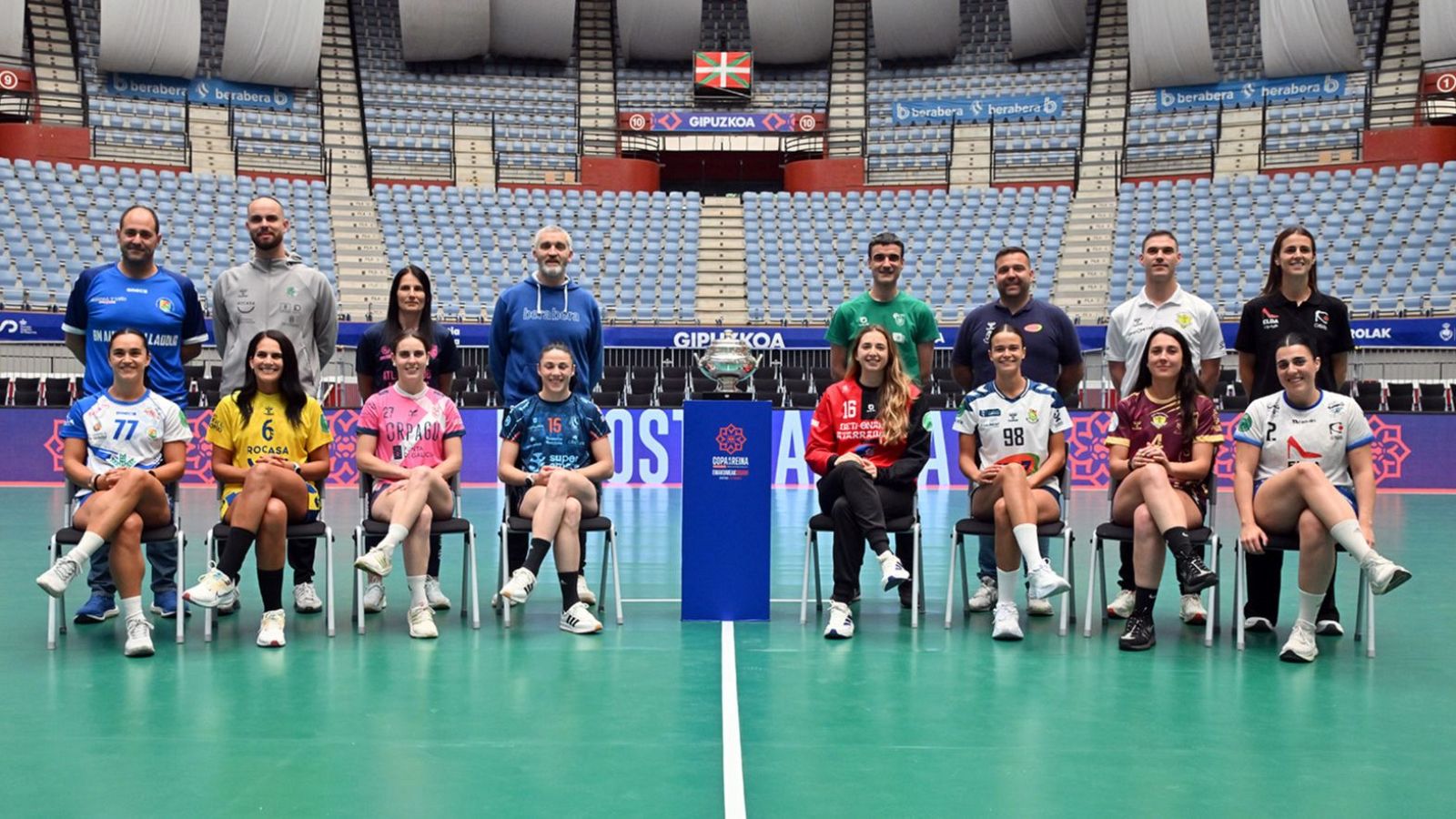 Fotografía de grupo de jugadoras y entrenadores de balonmano con el trofeo de la Copa de la Reina en un pabellón deportivo.