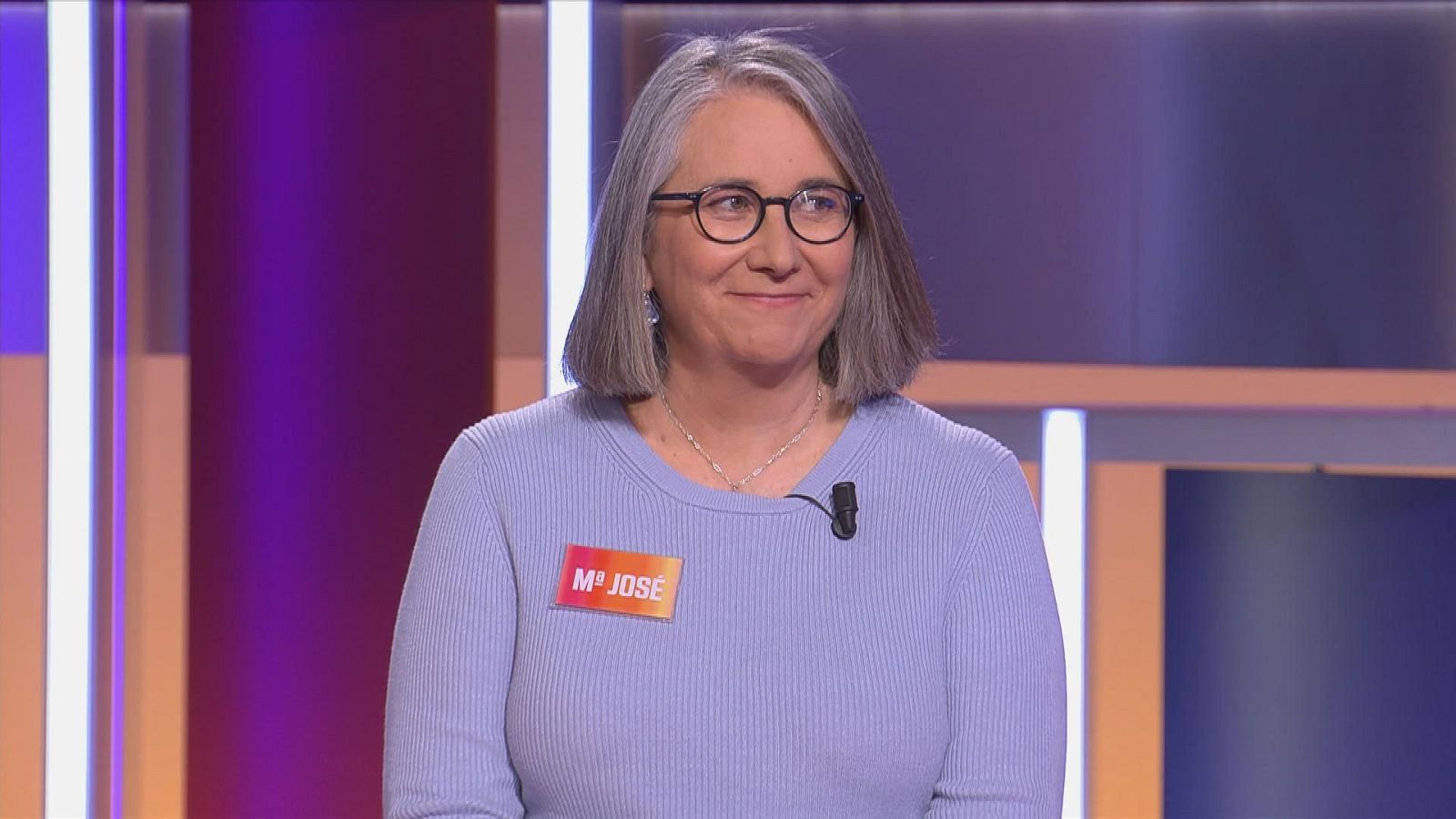 María José Cano, concursante de 'Saber y Ganar', sonríe a la cámara con gafas redondas, jersey lila y chapa naranja 'Mª JOSÉ'. Fondo de estudio de televisión con luces moradas y azules.