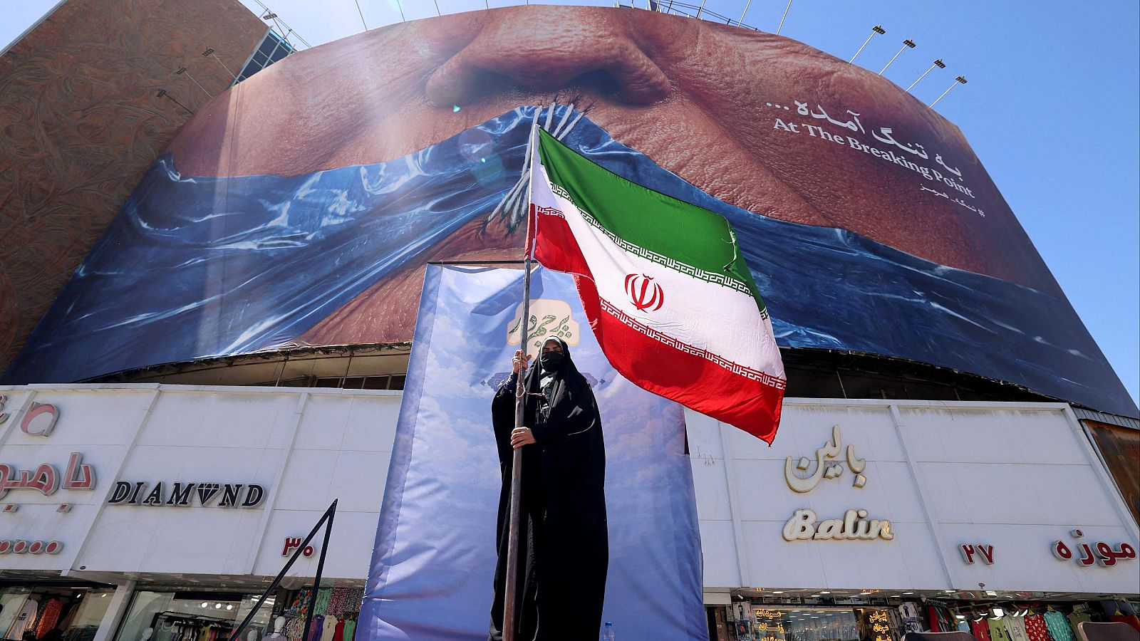 Mujer con chador sostiene bandera iraní frente a valla publicitaria con rostro de Trump, agua turbulenta y texto "At The Breaking Point." en inglés y persa.