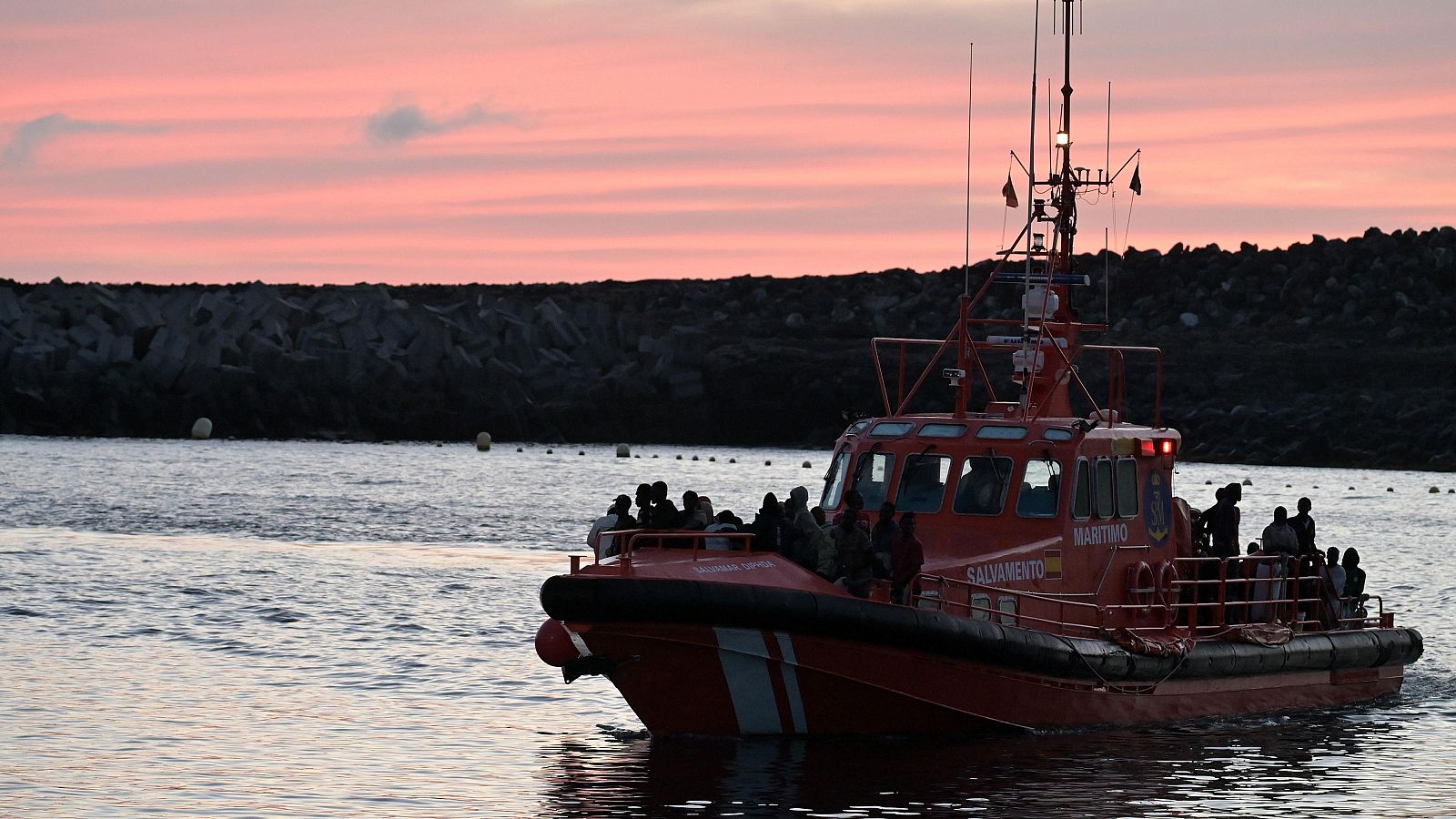 Embarcación de Salvamento Marítimo roja con "SALVAMENTO MARÍTIMO" visible, transporta a numerosas personas como siluetas, con un cielo crepuscular y costa rocosa al fondo.