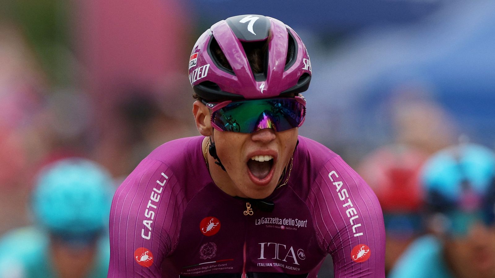 Primer plano de Paul Magnier celebrando, con casco magenta "SPECIALIZED", gafas iridiscentes y maillot "CASTELLI" con logos "La Gazzetta dello Sport" e "ITA".